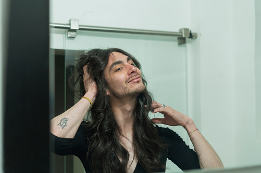 A middle-aged Hispanic man in his 30s with long black hair stands in front of the bathroom mirror at home, caressing his face as he blurs the makeup on his face