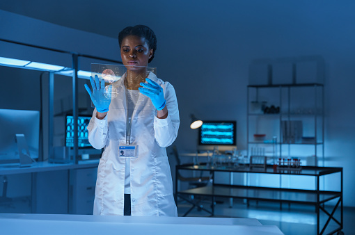 A young serious African - American doctor in a lab coat works with a small HUD screen in a modern laboratory