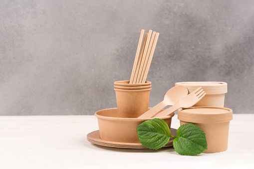 Eco paper utensils - paper plates, food containers, cups, drinking straws and wooden cutlery set against gray wall background with copy space. Sustainable food packaging concept. Eco tableware