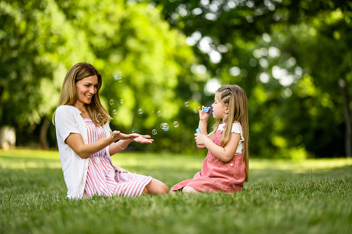 Happy mother and little daughter making soap bubble while having fun in nature on sunny spring day.