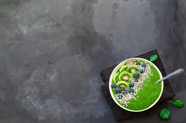 Breakfast green smoothie bowl with kiwi, blueberries and sunflower seeds on gray concrete background. Top view with space for text.
