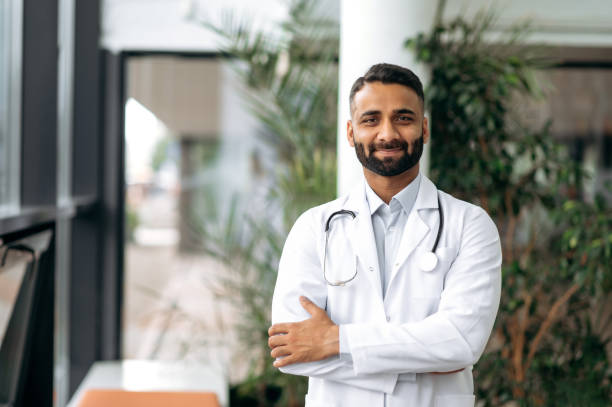 Portrait of handsome smart indian professional therapist in medical uniform and stethoscope, standing in hospital on blurred background looking at camera and smiling friendly stock photo