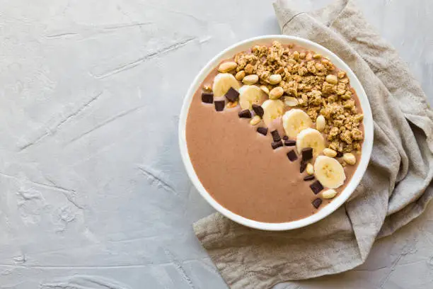 Chocolate smoothie bowl with bananas, granola and peanuts on light gray concrete background. Healthy vegetarian breakfast. Top view.