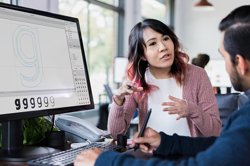 A female design professional gestures as she discusses an idea with a male colleague. The design is seen on a computer monitor.