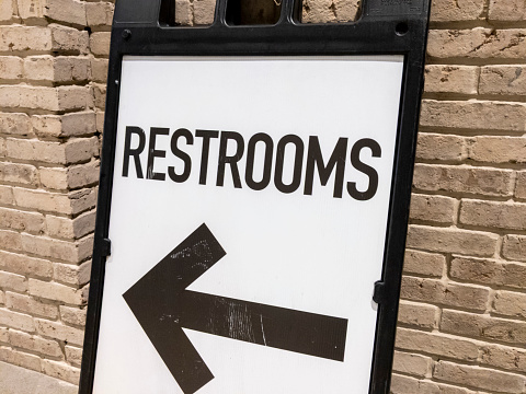 Angled view of a sandwich board pointing the direction of public restrooms, leaning against a brick wall