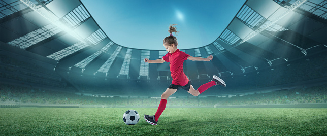 Sportive girl, football player in red uniform in motion, hitting ball at the open air stadium. Championship. Concept of sport, competition, movement, lifestyle, childhood