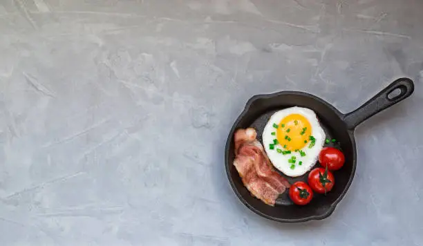 Fried egg with bacon and cherry tomatoes in iron skillet on light gray concrete background. Top view with space for text.