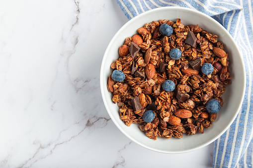 Homemade chocolate granola, muesli with almonds, hazelnuts and blueberries in bowl on white marble background. Healthy breakfast. Top view. Flat lay minimalist design. Space for text.