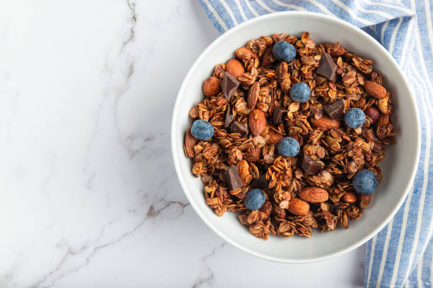 Chocolate granola, muesli with almonds, hazelnuts and blueberries Homemade chocolate granola, muesli with almonds, hazelnuts and blueberries in bowl on white marble background. Healthy breakfast. Top view. Flat lay minimalist design. Space for text. granola stock pictures, royalty-free photos & images