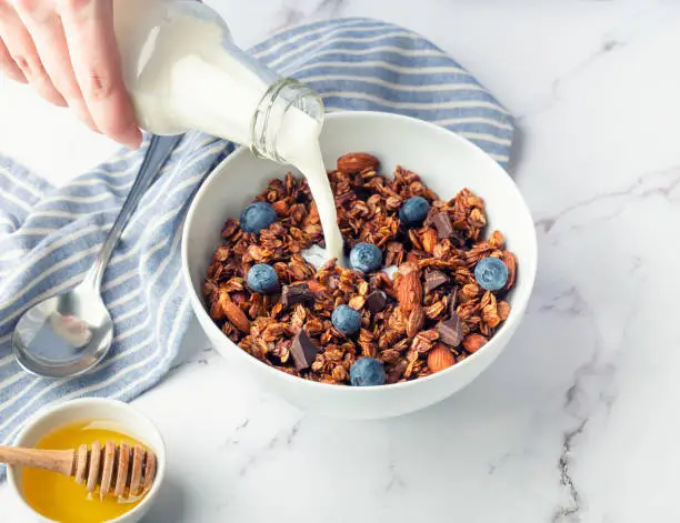 Homemade granola, muesli with pieces of dark chocolate, nuts, blueberries and pouring milk in bowl on white marble background. Healthy breakfast.