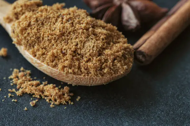 Natural brown sugar or unrefined sugar on wood spoon and cinnamon stick and anise on black granite table with copy space. Sweet seasoning for good health. Ingredient prepared for cooking or bakery.