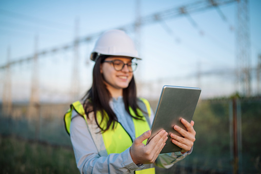 Young female engineer with protective headwear smiling and using digital tablet at power plant.