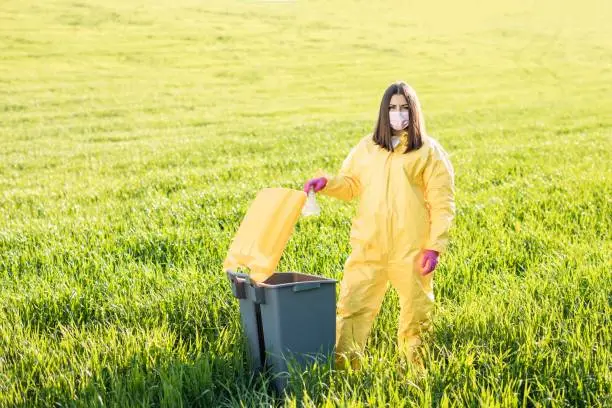 Photo of A woman in a yellow protective suit stands in the middle of a green field and holds a plastic bottle in her hands to throw in the tank