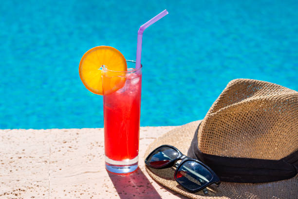 un cóctel rojo en una copa con una rodaja de naranja, un sombrero de hombre para el sol y gafas de sol en el borde de la piscina del hotel. - swimming pool resort swimming pool poolside sea fotografías e imágenes de stock