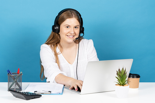 Young telephone or telemarketing operator working on laptop. Blonde woman looking at camera isolated on blue background.