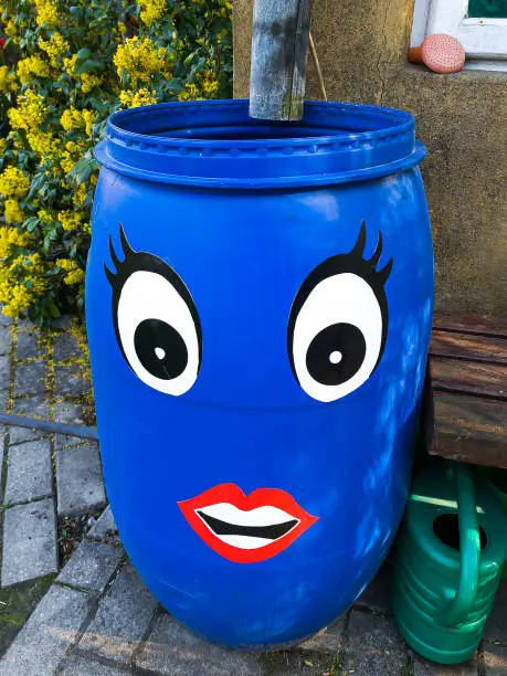 Photo of A blue barrel is attached to the house by the gutter. Eyes and mouth are glued to the barrel.