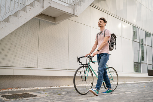 One man young adult male with brown hair and mustaches walking by the building with rucksack on his back and bicycle happy smile joyful real people copy space side view full length