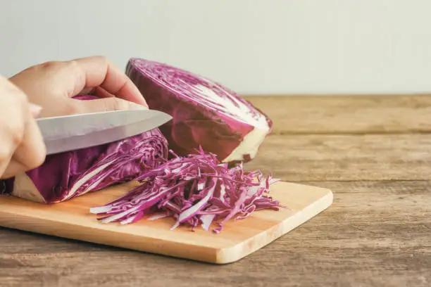 Woman use knife kitchen to chopped or sliced fresh purple cabbage on cutting board to shredded on wood table. Prepare vegetable for cooking cabbage salad or coleslaw. Homemade food concept.