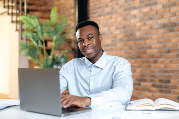 istock Positive confident black man using laptop in modern office. Portrait successful businessman, freelancer, manager, in formal shirt, working and typing on laptop, looking at camera, smiling friendly 1402203758