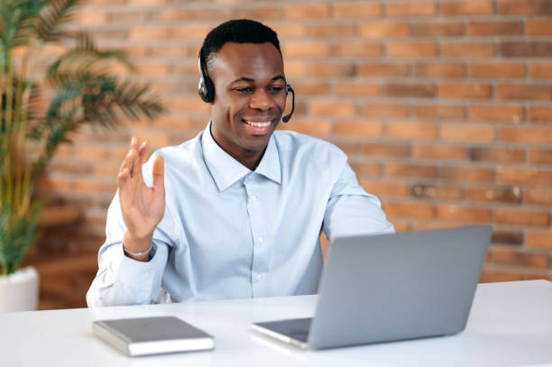 istock Positive attractive African American guy in headphones, hotline operator, support worker, online consultant sits at a laptop in the office, works with a client on video call using headset, greetings 1402203786
