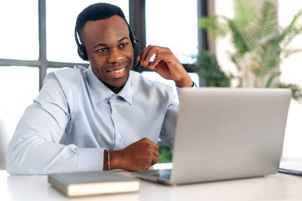 istock Successful pleasant African American guy in headphones, hotline operator, support worker, online consultant is sitting at a laptop in the office, talking to a client on a video call, smiling friendly 1402203792