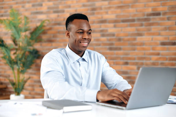 istock Positive confident black man using laptop in modern office. Successful businessman, freelancer, manager, in formal shirt, browsing email, working and typing on laptop, working on a project, smiling 1402203796