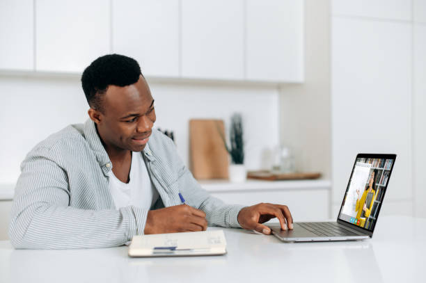 istock Distance online learning. African American smart guy, student or freelancer, sits at a table at home in the kitchen, has an online lecture, takes notes, on the screen female teacher teaches remotely 1402203862