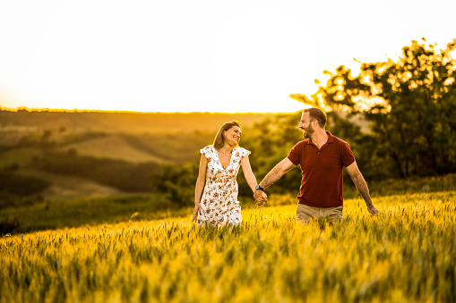 Cheerful couple holding hands while walking outdoors on a field during sunset.