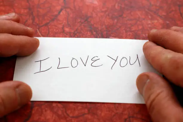 I love you printed in black on a white piece of paper with white male fingers on each side of the paper. White male fingers touching a white index card with the message I LOVE YOU written on it.