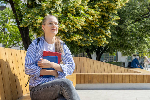 istock Female student holding books when sitting on a park bench 1402233191