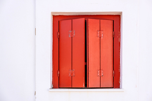 Window with semi closed wooden shutters red color on white wall background. Cyclades island house front view, Greek traditional architecture.