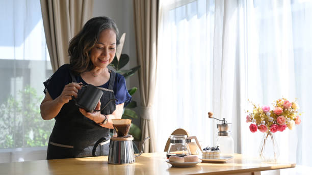 istock Positive middle aged standing at kitchen table, brewing drip coffee and enjoys her morning. 1402256745