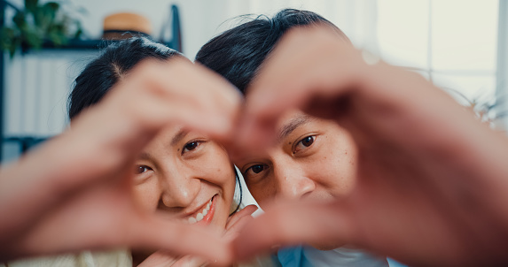 Close-up Young Asian married couple with casual sitting on couch make hand to be love sign heart shape and looking at camera with sweet and happy moment in living room at home.