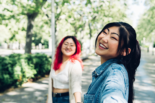Happy young Japanese female in denim shirt smiling and taking selfie on sunlit park alley while holding hand of girlfriend on weekend day