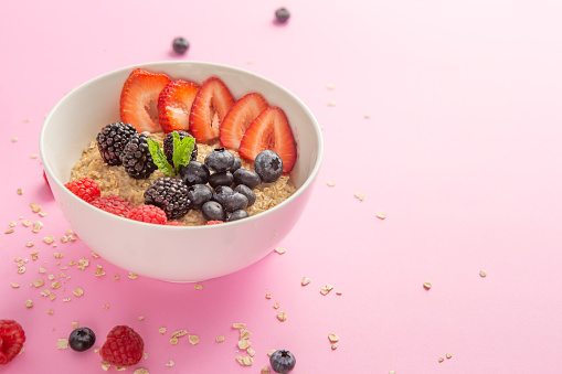 Oatmeal bowl with mixed berries on pink background with scattering oat flakes .