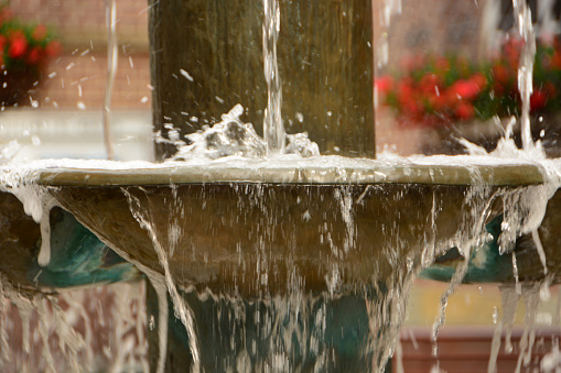 Fountain water flowing over the edge of the storage tank. In the background out of focus red blooming flowers.