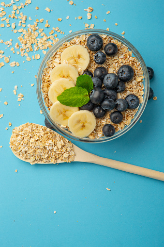 Oatmeal flakes bowl with banana and blue berries on blue background.