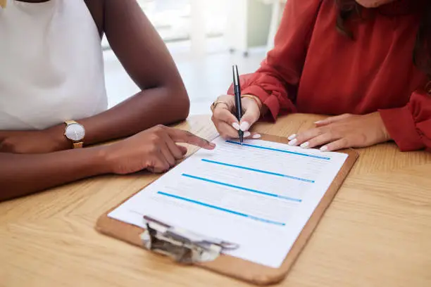 Photo of Two unknown ethnic business woman sitting together and signing office contract. African american professional using hand gesture to point on paperwork. Mixed race colleague agreeing to document deal