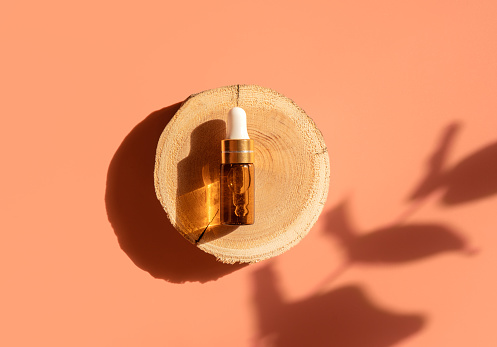 Amber glass dropper bottle on woodcut in the sunlight with eucalyptus flower shadows. Top view. Luxury and natural cosmetics presentation. Testers, beauty samples perfumery concept. Shades and lights