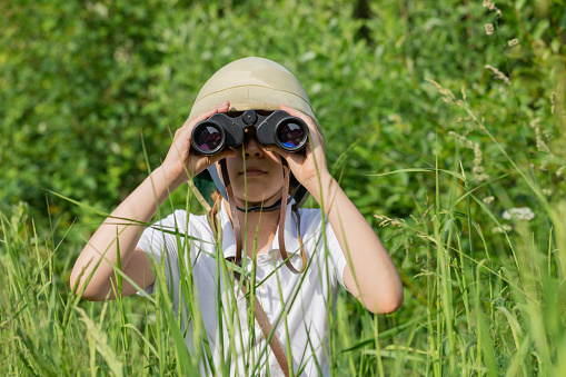 Preteen girl wearing pith helmet hiding in the grass looking through binoculars observing summer nature. Discovery and adventures concept