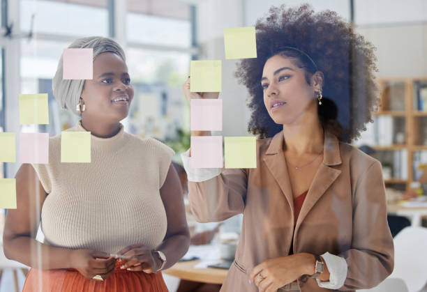 istock Mixed race businesswoman with afro using visual aid to brainstorm in the office. Hispanic professional standing and showing african american colleague. Sharing ideas and planning marketing strategy 1402295043