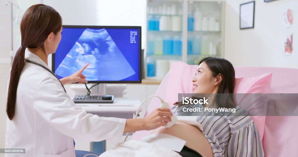 Prenatal exam concept back view of female asian doctor wear white coat uses ultrasound machine helps young pregnant woman on bed do prenatal exam at clinic Taiwan Stock Photo