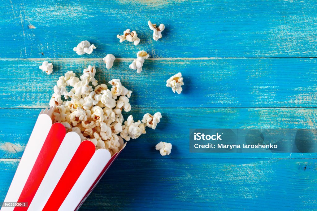 Fresh popcorn spilled from a cardboard striped cup on a blue wooden background with copy space. Top view. Popcorn Stock Photo