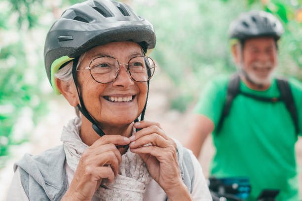 남편이 웃고 있는 가운데 야외에서 자전거를 타고 자연을 즐기며 웃고 있는 한 노인의 초상화. 건강한 느낌의 안경을 가진 성숙한 여성의 헤드 샷. 시니어는 헬멧을 착용하고 자전거로 여행합니 - action mature couple senior adult mature adult 뉴스 사진 이미지