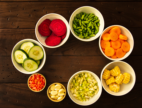 High angle view of fresh vegetables in bowls as cucumber, tomato, garlic clove, celery chopped, corn, carrot, green bean and beet.  in half or chopped up vegetables used as healthy food ingredients to cook.