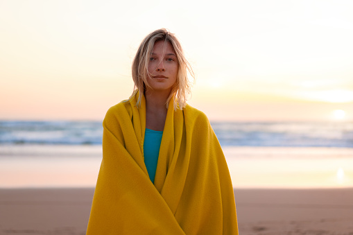 Beautiful young woman wrapped in yellow blanket standing on sandy beach at sunset looking camera. Beautiful caucasian female person wrapped cozy plaid standing alone on ocean beach in dusk summer time