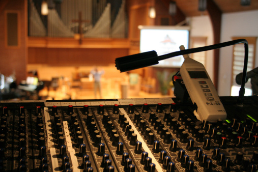 Peaceful view of a church sanctuary and worship service in soft focus behind a view of a sound/audio mixing board (console) and sound level meter.  Projection screen and speaker/preacher in background, with organ pipes and cross showing that it is a church.