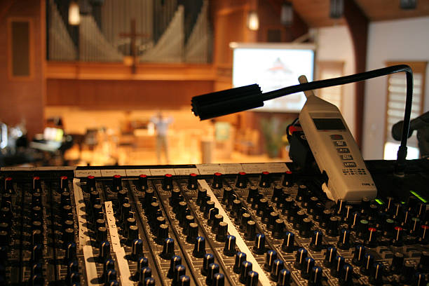 Running sound at a church worship service Peaceful view of a church sanctuary and worship service in soft focus behind a view of a sound/audio mixing board (console) and sound level meter.  Projection screen and speaker/preacher in background, with organ pipes and cross showing that it is a church. place of worship stock pictures, royalty-free photos & images