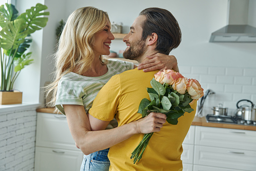 Cheerful young man surprises his girlfriend with bunch of flowers at the kitchen