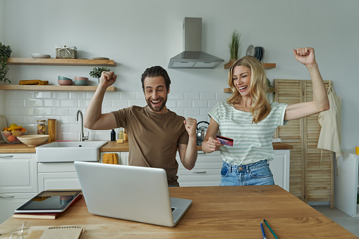 Happy young couple keeping arms raised while shopping online from domestic kitchen